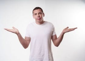 Adult man in white t-shirt shrugs in studio, expressing confusion against a plain background.
